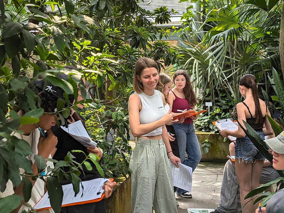 Das Foto zeigt einige Schülerinnen und Schüler zusammen mit einer Studentin, die sie anleitet, während des Rollenspiels an einer Station des Workshops. Die Station befindet sich im Tropenschauhaus des Botanischen Gartens.